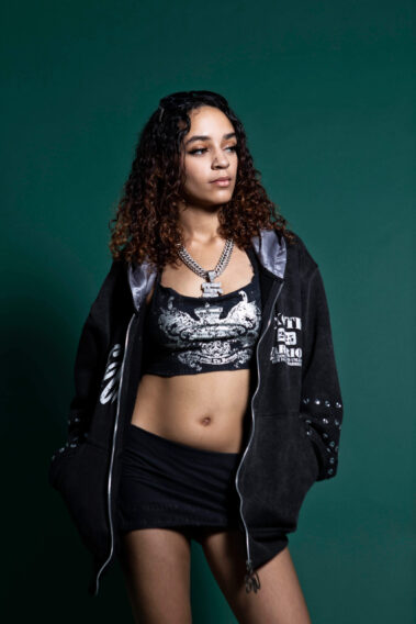 Model in oversized black streetwear posing against a green backdrop during the MAYHEM event.