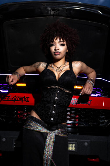 Model in black fashion outfit leaning against a customized truck with colorful lighting at night.