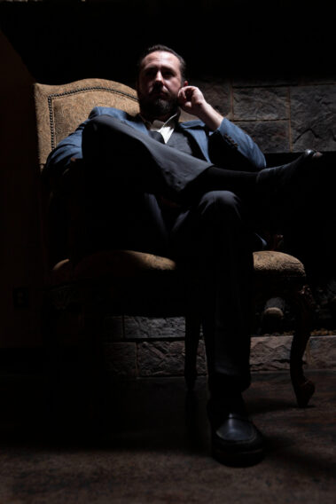 Low-key cinematic portrait of a man in a blue suit seated in an ornate chair beside a stone fireplace during a 1940s-themed photomeet at Castle Falls.
