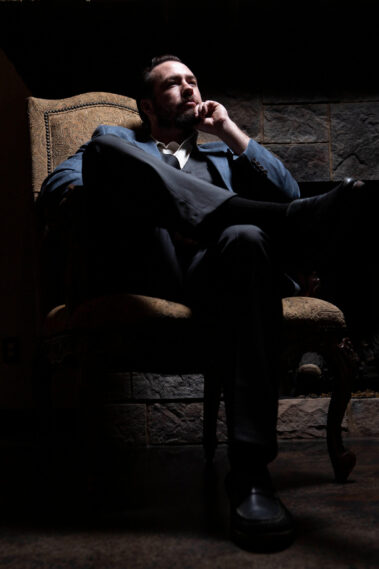 Portrait of a man in a blue suit seated thoughtfully in a vintage chair with dramatic lighting during a 1940s-inspired photography event.