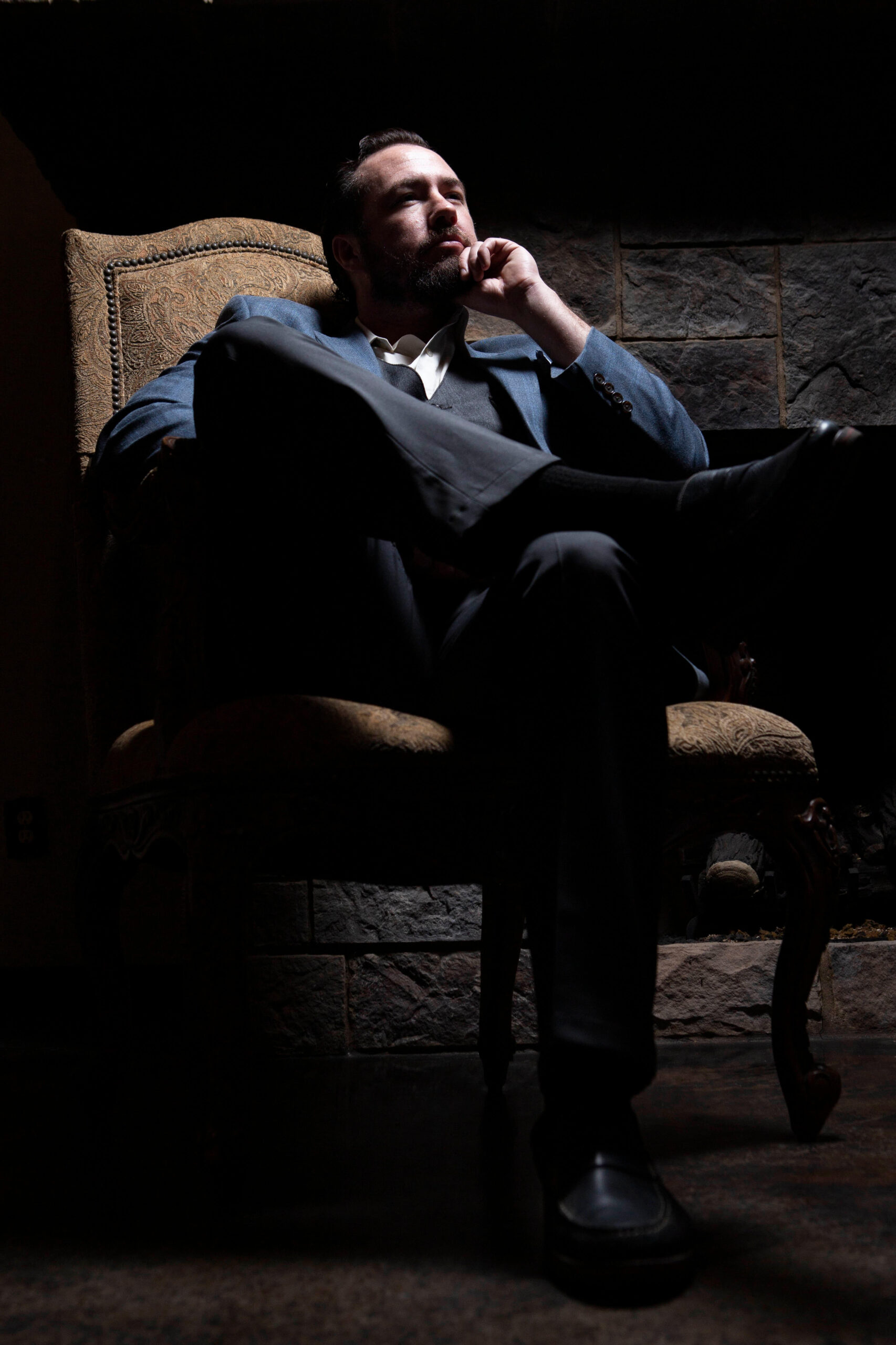 Portrait of a man in a blue suit seated thoughtfully in a vintage chair with dramatic lighting during a 1940s-inspired photography event.