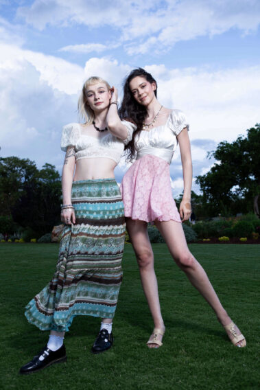 Models madisxnariana and caitlinrmathews posing together outdoors on a grassy garden lawn beneath a dramatic cloudy evening sky.
