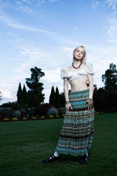 madisxnariana posing on the lawn at Will Rogers Gardens wearing a patterned skirt and white lace top beneath a soft evening sky.