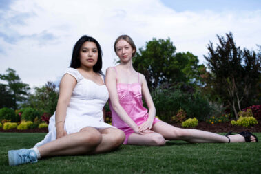 Alise_Rojo in a white dress seated beside Mariah_Rain_ in a pink satin dress on the grass during an outdoor garden portrait session.