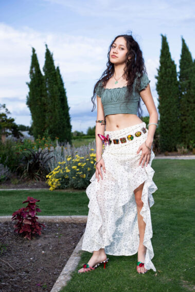 jadyntrann standing among colorful flowers and tall trees in a flowing white layered skirt and green crop top during an outdoor fashion photoshoot.