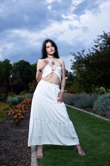 Bella Bazan posing in a white two-piece outfit along a garden pathway beneath dramatic evening clouds during an outdoor photoshoot.