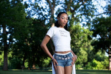 Fashion portrait of a model in a patterned skirt and layered jewelry posing in a grassy park surrounded by large trees.