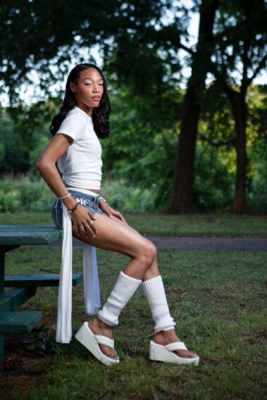 Fashion portrait of a model wearing a white "Protect the Dolls" shirt and denim shorts in a park.