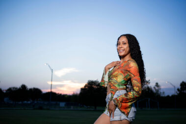 Portrait of a model in a patterned dress standing in a field at sunset during an outdoor photography event.