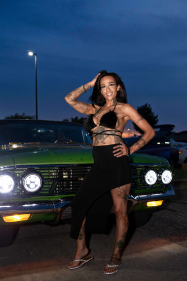 A tattooed model poses in front of a green classic Chevrolet at dusk.