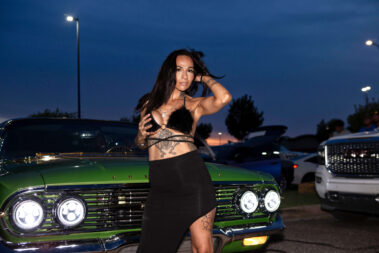 A model poses beside a green classic car illuminated by bright headlights at a nighttime car show.