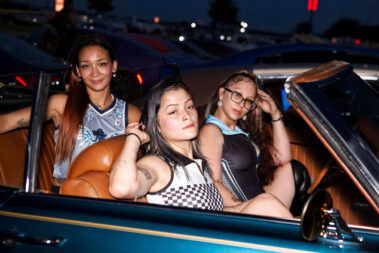 Three Ice Angels models sit inside a blue convertible during a nighttime car show.