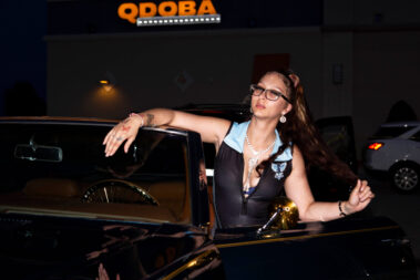A promotional model poses beside a classic black vehicle during an evening car meet.