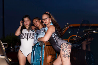 Three promotional models pose beside a classic blue convertible during a nighttime car show.