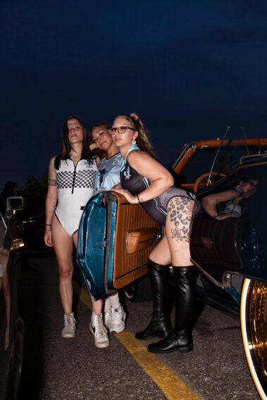 Three models pose beside a classic convertible parked in a car show lot at dusk.