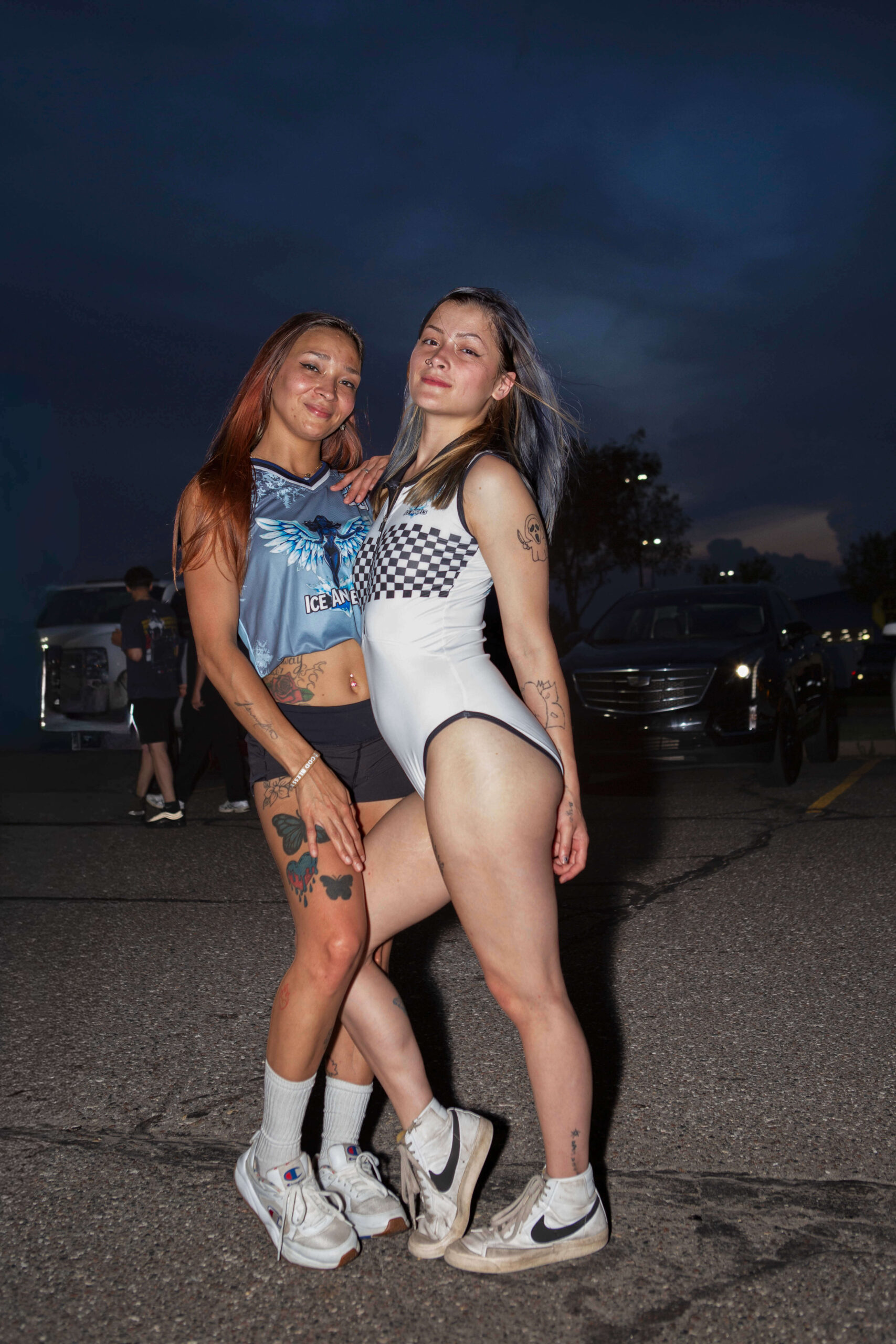 Ice Angels Under the Storm Two Ice Angels models pose together in a parking lot at dusk during the Street Motion car show as storm clouds gather overhead.