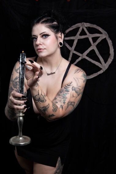 Gothic model Kari Anne holds a burning black candle in a silver candlestick, carefully positioning her fingers near the flame. Her tattooed arms and black dress contrast with a dark backdrop featuring a large pentagram.
