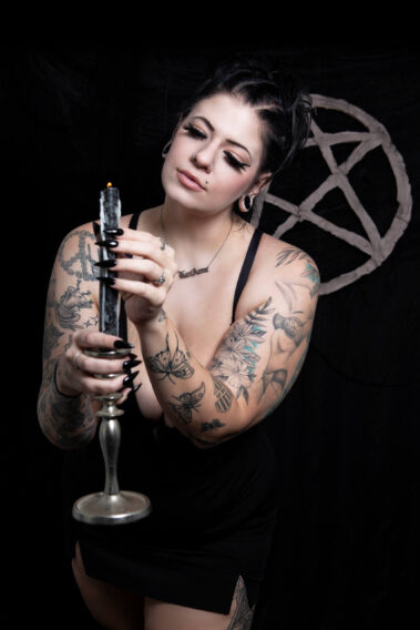 Kari Anne gazes downward at a burning black candle while holding an antique silver candlestick. Her tattooed arms, dramatic black nails, and black dress stand out against a dark background featuring a pentagram.