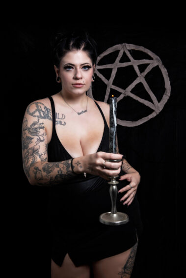 Kari Anne faces the camera while extending a silver candlestick holding a burning black candle. She wears a black dress, and her tattooed arms are illuminated against a dark studio backdrop featuring a pentagram.