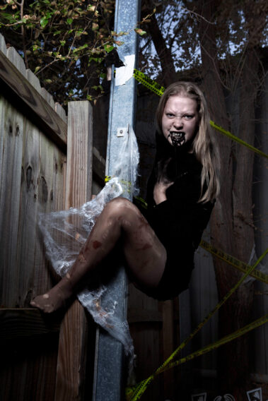 Blonde Halloween model wearing a distressed black dress, dark gloves, theatrical fangs, and black mouth makeup poses aggressively against a metal post surrounded by spiderwebs and crime scene tape.