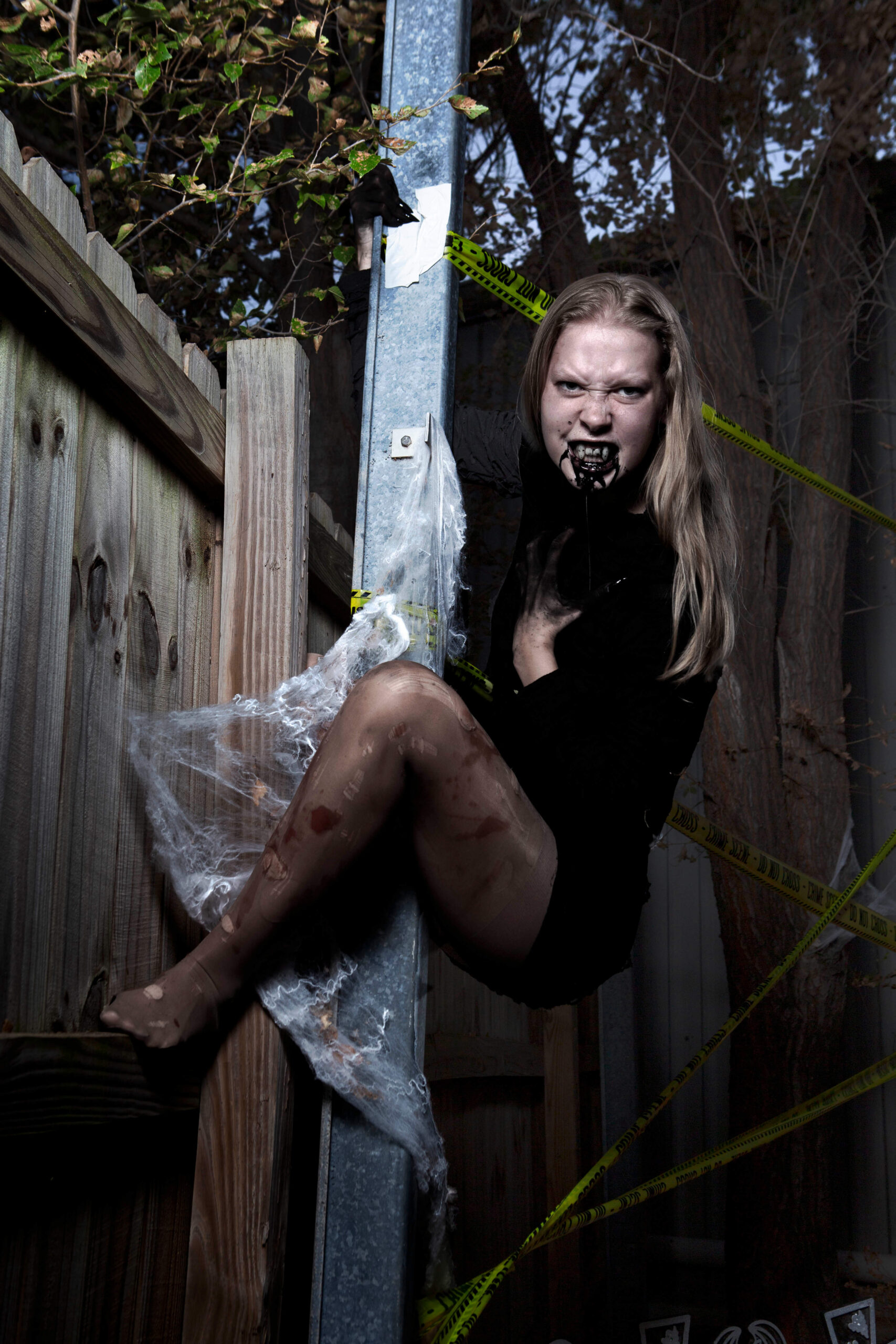 Blonde Halloween model wearing a distressed black dress, dark gloves, theatrical fangs, and black mouth makeup poses aggressively against a metal post surrounded by spiderwebs and crime scene tape.