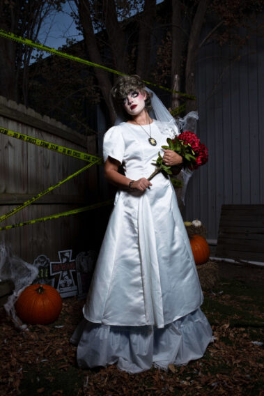 Gothic bride in a white wedding gown and veil holds a bouquet of deep red flowers while standing among pumpkins, fallen leaves, artificial spiderwebs, and yellow crime scene tape.