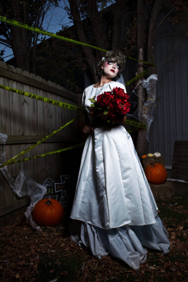 Model dressed as a ghostly bride in a white wedding gown and veil holds a large bouquet of dark red flowers while posing in a Halloween graveyard setting with pumpkins and crime scene tape.