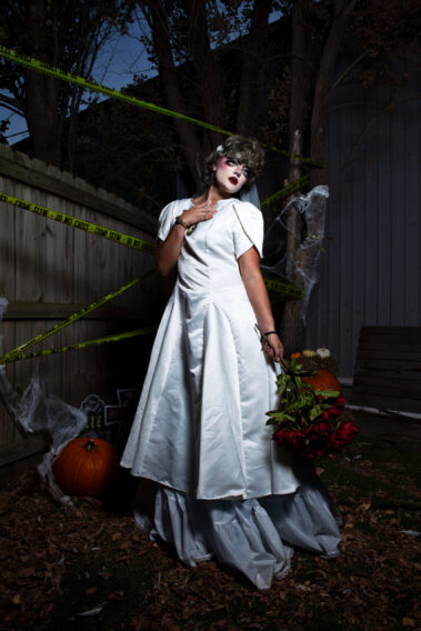 Halloween model dressed as a ghostly bride in a white gown stands outdoors holding a bouquet of red flowers at her side, surrounded by pumpkins, fallen leaves, and crime scene tape.