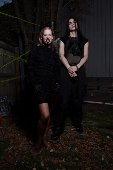 Two gothic Halloween models pose together outdoors, one with blonde hair, theatrical fangs, and black mouth makeup, and the other with long black hair, dark clothing, and silver jewelry.