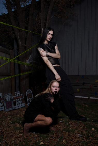Long-haired gothic model dressed in black stands behind a crouching blonde Halloween model wearing theatrical fangs and dark makeup in an outdoor graveyard-themed setting.