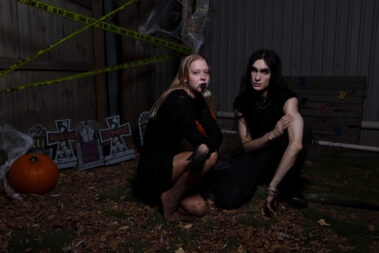 Two gothic Halloween models crouch together outdoors among fallen leaves, pumpkins, decorative gravestones, and yellow crime scene tape.