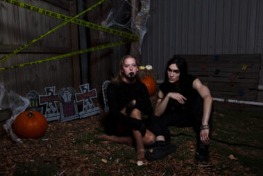 Blonde Halloween model wearing theatrical vampire fangs and black mouth makeup pretends to bite the neck of a long-haired gothic model dressed in black.