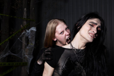 Long-haired gothic model dressed in black kneels beside an ornate skull-decorated candelabra with three dark candles against a backdrop of dense artificial spiderwebs.