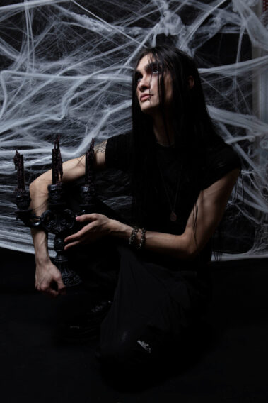 Gothic model with long black hair and dark clothing kneels while holding a black skull-decorated candelabra and looking upward against an elaborate white spiderweb backdrop.