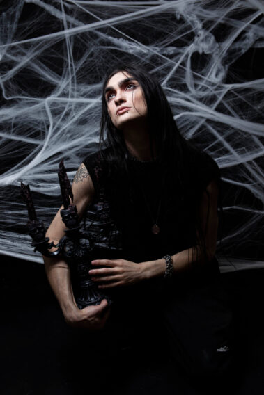 Long-haired gothic model wearing black clothing and silver jewelry kneels while holding a three-candle black candelabra beneath a large white spiderweb installation.