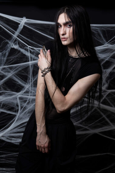 Gothic model with long black hair, dark clothing, silver jewelry, and crossed arms poses against a backdrop of white spiderwebs at CDR Studios.