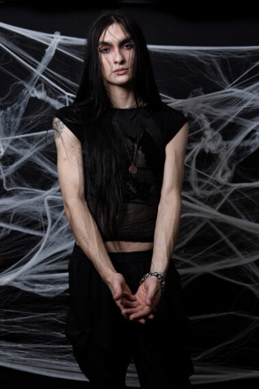 Long-haired gothic model wearing a black sheer top and silver jewelry stands facing the camera with hands clasped against an elaborate spiderweb backdrop.