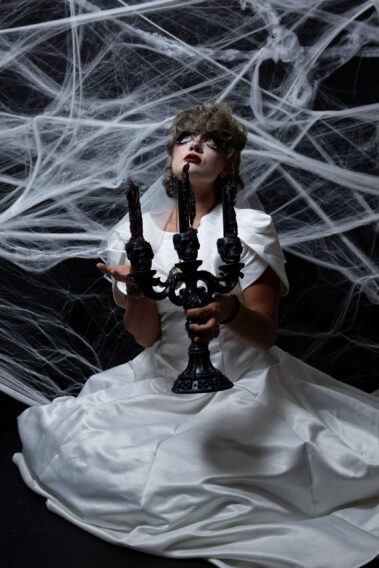 Model wearing a white bridal gown, veil, and dramatic Halloween makeup holds an ornate black three-candle candelabra beneath a large spiderweb backdrop.