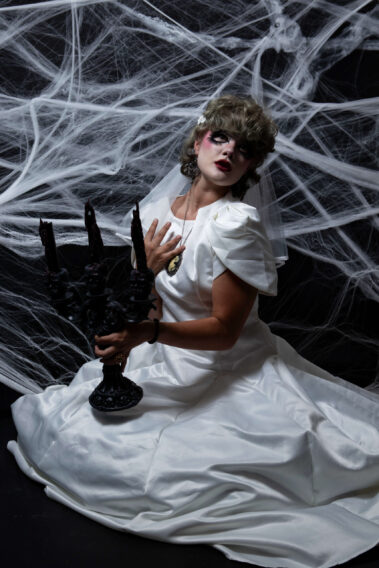 Gothic bride in a white wedding dress and veil poses with one hand against her chest while holding a black skull-decorated candelabra in front of white spiderwebs.