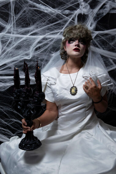 Model dressed as a gothic bride in a white satin gown and veil holds a black skull candelabra with partially lit candles against a dense spiderweb backdrop.