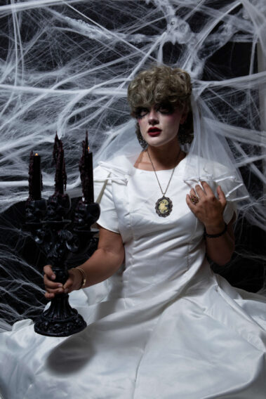Gothic bride wearing a white satin wedding dress and veil holds a black three-candle candelabra while looking toward the camera against a spiderweb-covered background.