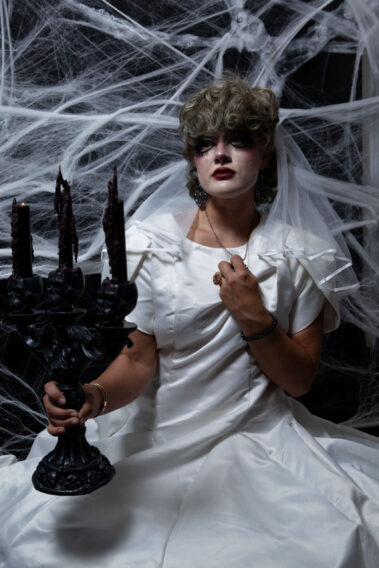 Close portrait of a gothic bride in a white wedding gown and veil holding an ornate black candelabra while clutching the fabric of her dress.